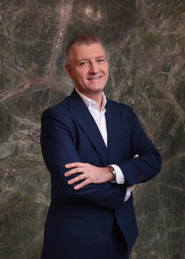 Bărbat în costum albastru și cămașă albă, zâmbind în fața unui fundal cu textură de piatră