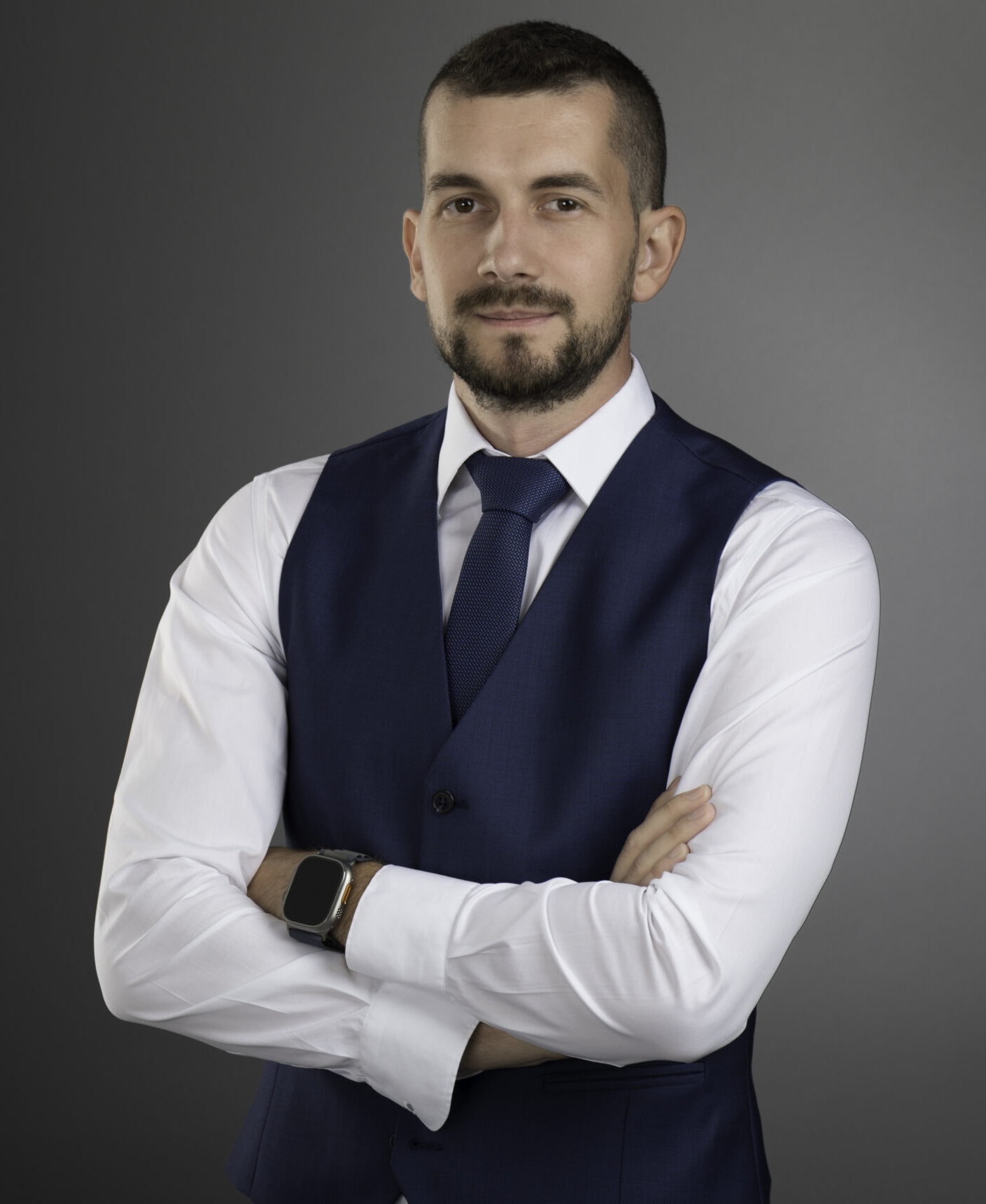 Portret profesional business al unui bărbat tânăr îmbrăcat în vestă bleumarin și cămașă albă, pe fundal gri gradient.