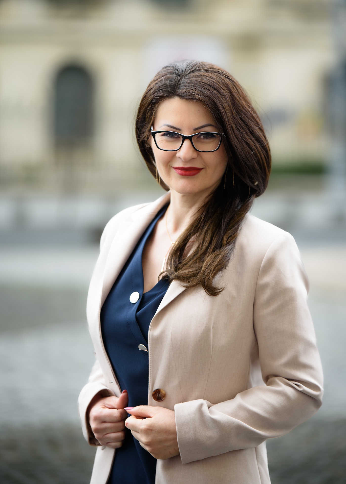 Femeie elegantă purtând ochelari și sacou bej, zâmbind în aer liber, în fundal urban blurat