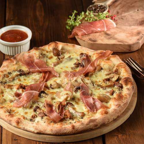 Pizza artizanală cu blat crocant, ciuperci și felii de prosciutto crudo, servită pe platou rustic din lemn, cu sos roșu alături